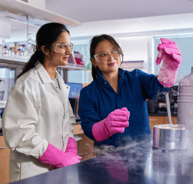 Two researchers wearing safety goggles and bright pink gloves work together in a laboratory. One researcher carefully handles equipment emitting cold vapor, while the other observes. Scientific instruments, tubing, and lab benches surround them.