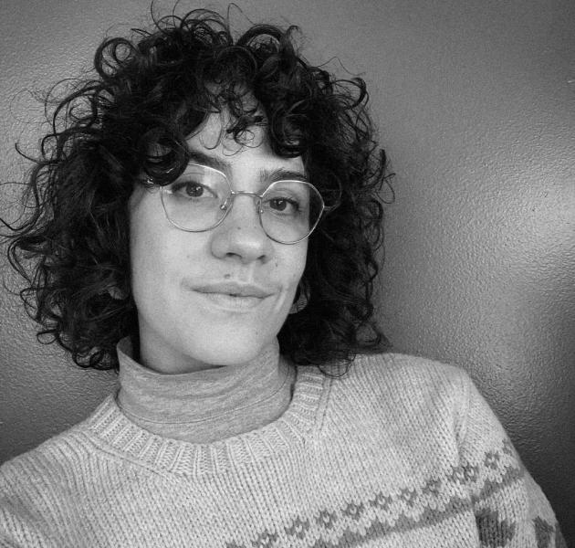 A black and white portrait of a person with curly dark hair and round glasses, wearing a turtleneck under a knit sweater, smiling faintly against a plain background.