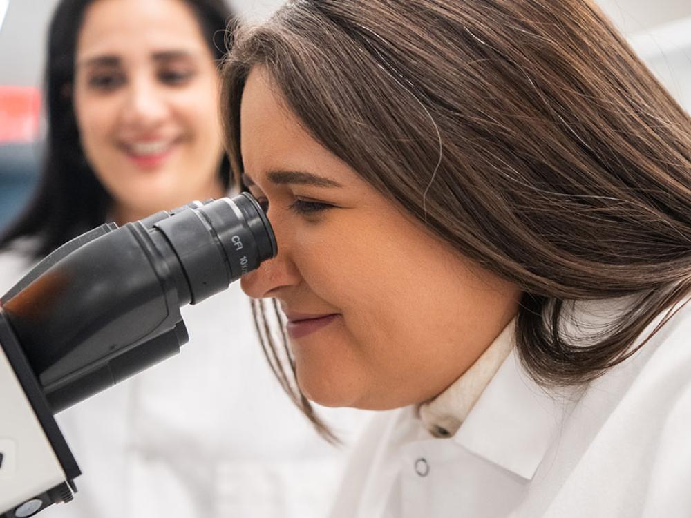 Person in a lab coat using a Nikon microscope, with another person smiling in the background.