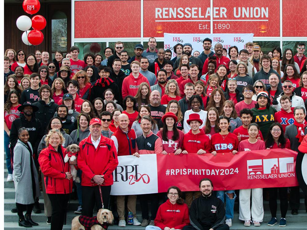 Group of people in red and black attire, holding banners with "RPI" and "#RPISPIRITDAY2024", standing in front of Rensselaer Union building, decorated with red and white balloons.